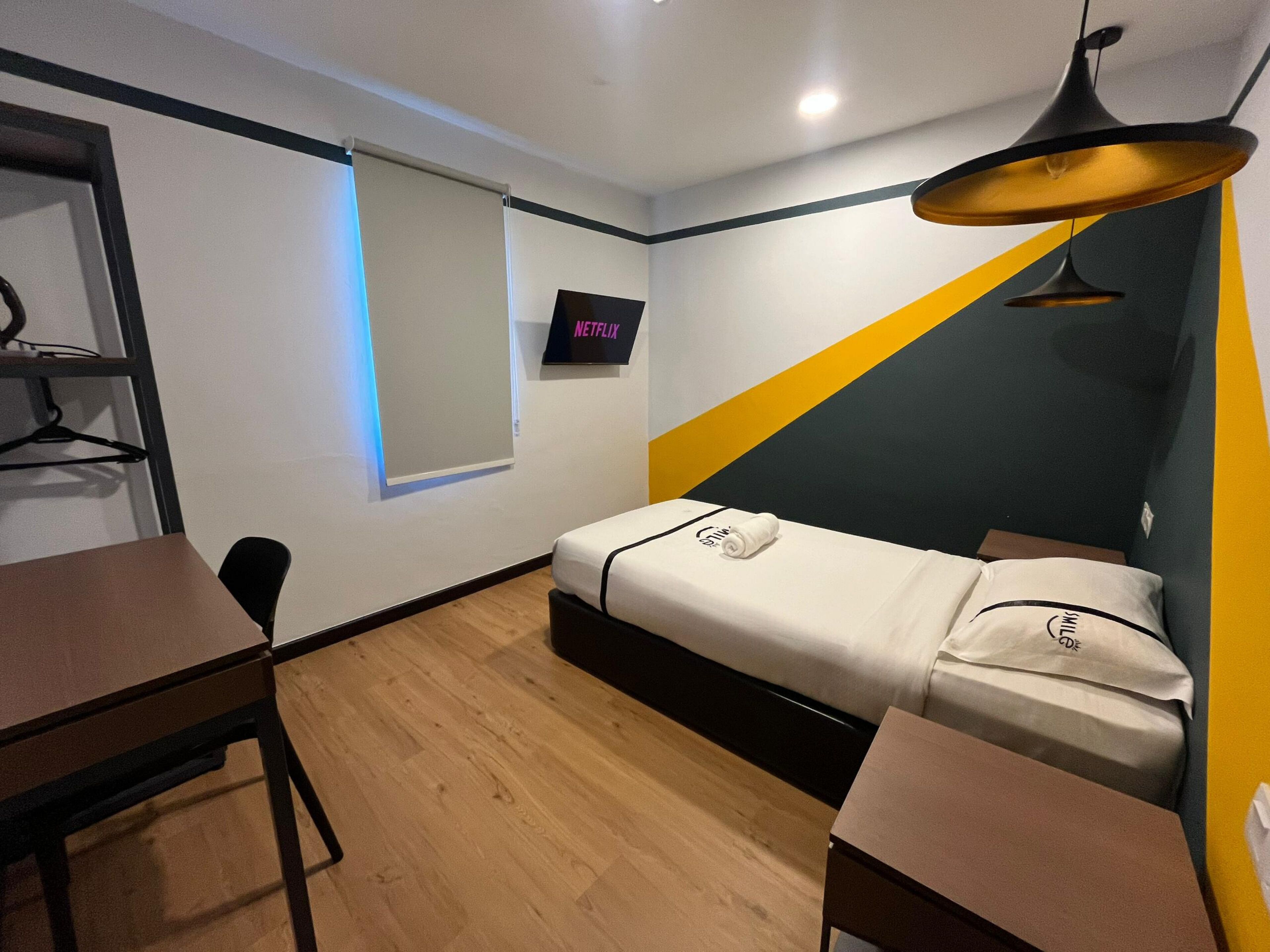 A small, modern room with a bed, a desk, a chair, and a TV screen displaying 'NETFLIX'.