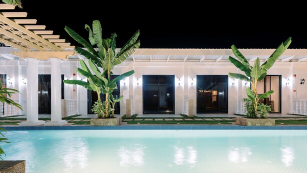 Un área de piscina con una pileta despejada, plantas en macetas y un edificio con columnas y puertas.