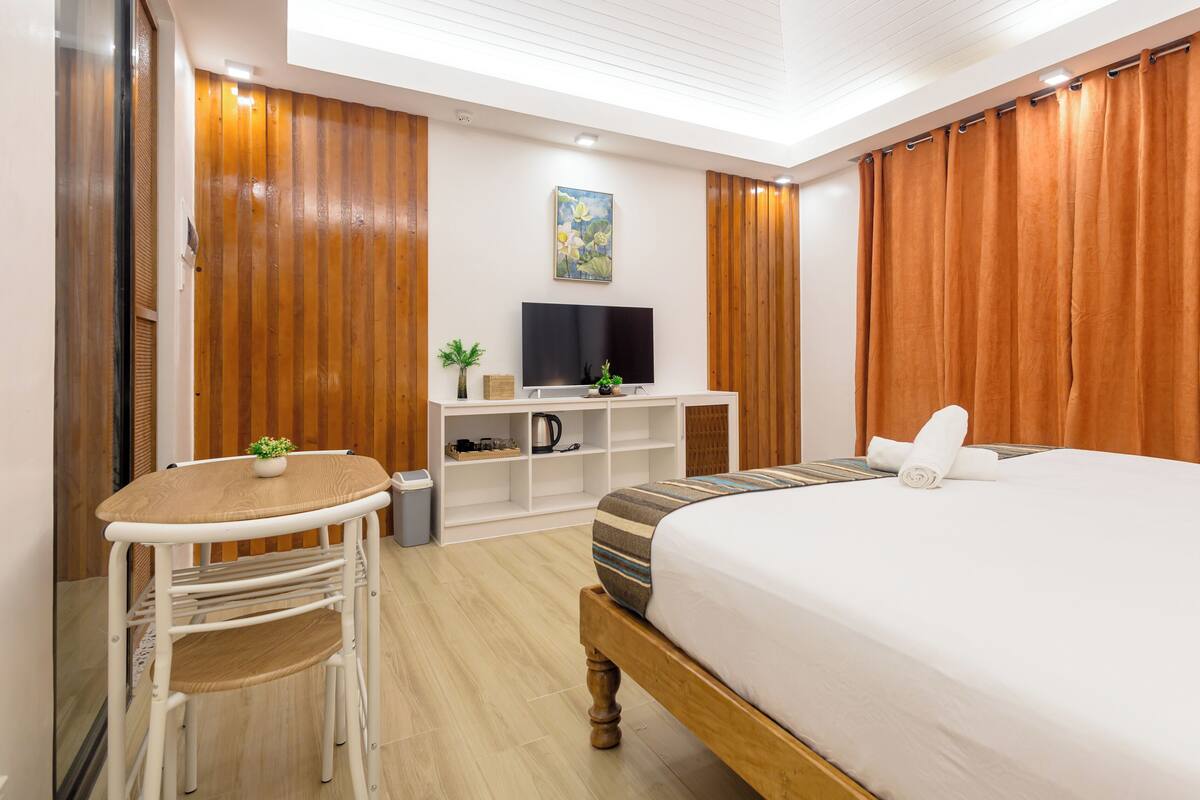 Hotellihuone, jossa on sänky, kirjoituspöytä, jossa on televisio, pieni pöytä tuoleineen ja puulattia.
