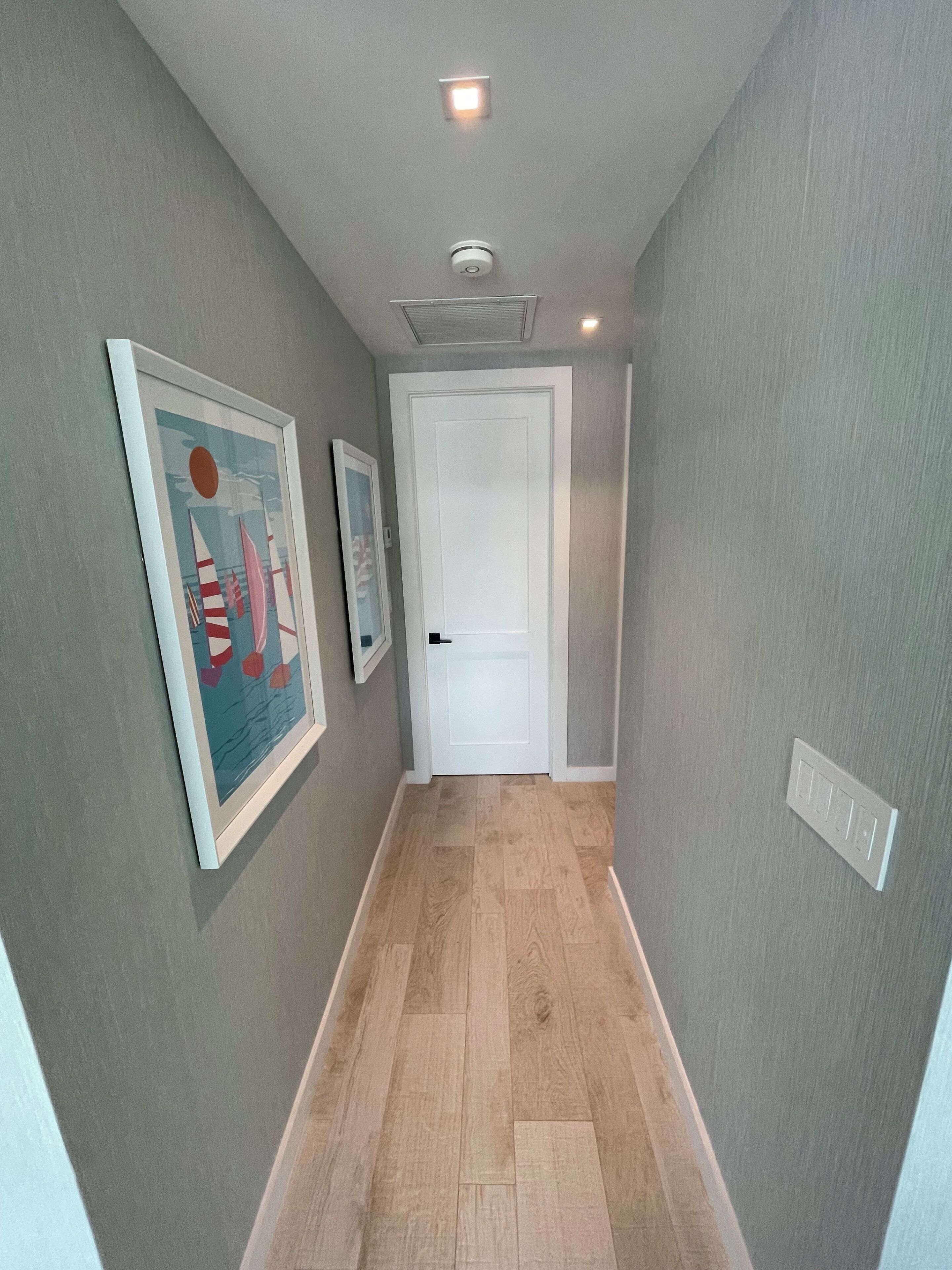 Un couloir avec un plancher en bois, une porte blanche au fond et deux tableaux encadrés accrochés au mur.