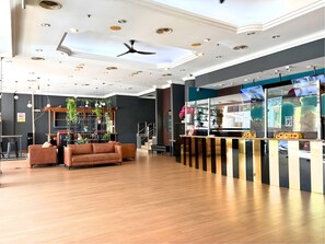 Eine moderne Hotelhalle mit Holzboden, einem braunen Sofa, einem Deckenventilator und einem Empfangsbereich mit Fernseher.