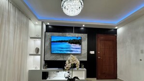 A modern living room with a flat-screen TV mounted on a marble wall, a glass coffee table, a wooden door, and a chandelier. - Casa Las América (Santo Domingo Este)