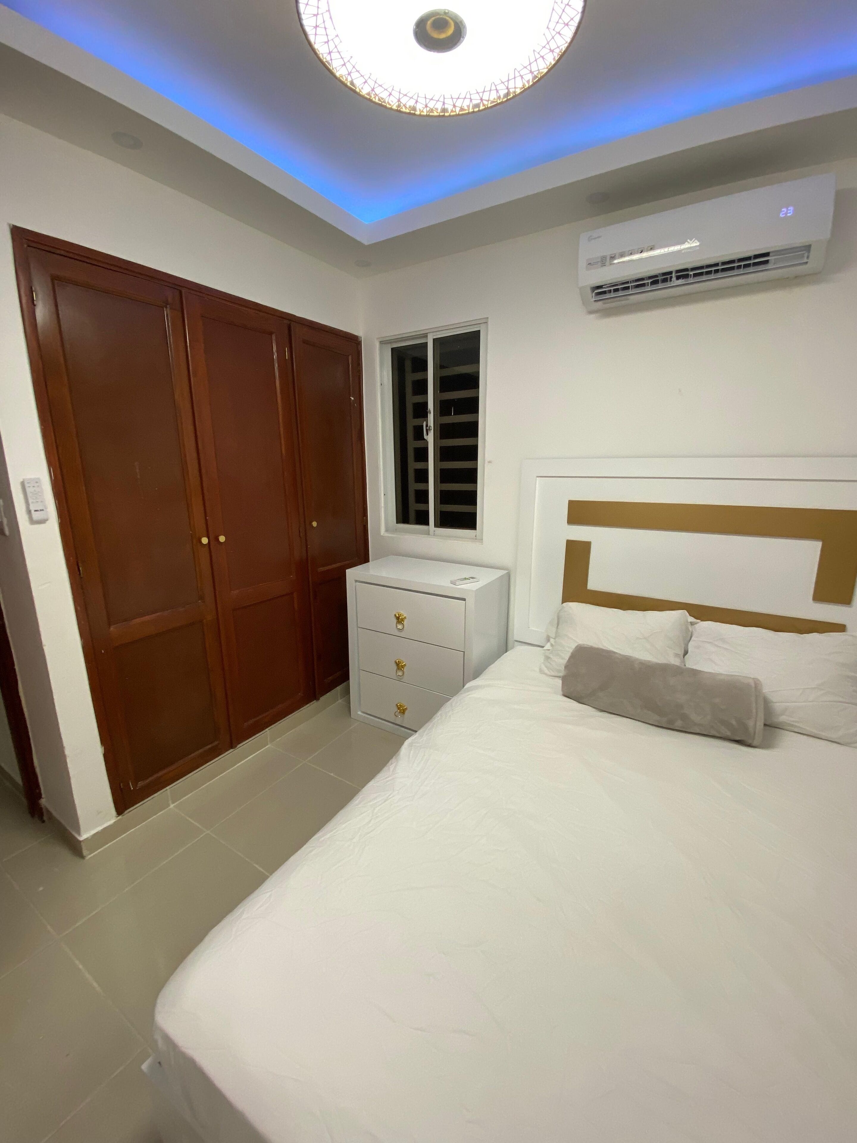 A bedroom with a bed, a wardrobe, a bedside table, an air conditioning unit, and a window.