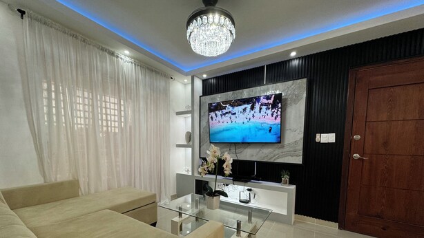 A modern living room with a beige sofa, a glass coffee table, a TV mounted on a marble wall, and a wooden door. - Casa Las América (Santo Domingo Este)