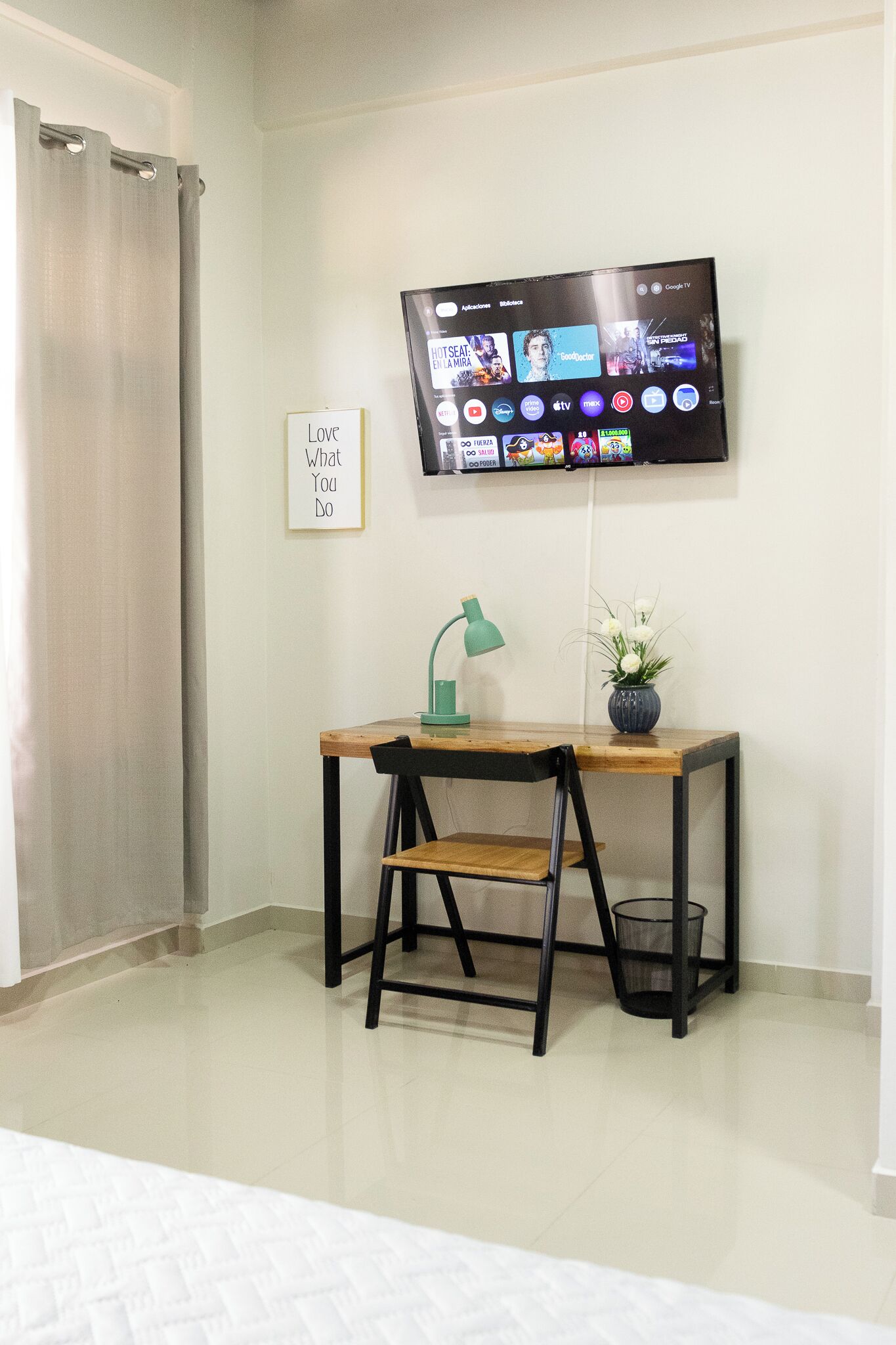 A hotel room with a wooden desk, a green desk lamp, a small potted plant, a TV mounted on the wall, and a sign that says "Love What You Do".