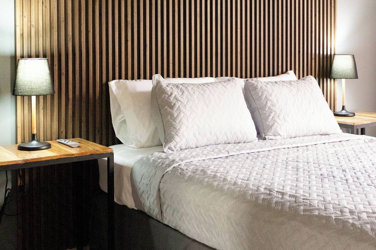 Een moderne slaapkamer met een houten lattenbovenstuk, een groot bed met witte beddengoed, twee nachtkastjes met lampen, en een klein nachtkastje.