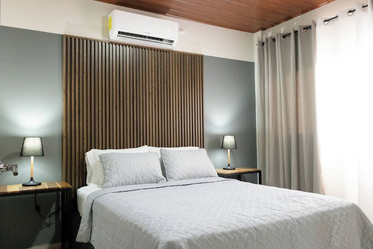 Een net opgemaakt bed met een grijze quilt, twee bedlampen, een houten lattenbalken hoofdbord, een airconditioning en een raam met gordijnen.