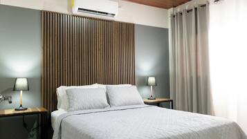 Una cama bien hecha con colcha gris, dos lámparas de noche, un cabecero de listones de madera, un equipo de aire acondicionado y una ventana con cortinas.