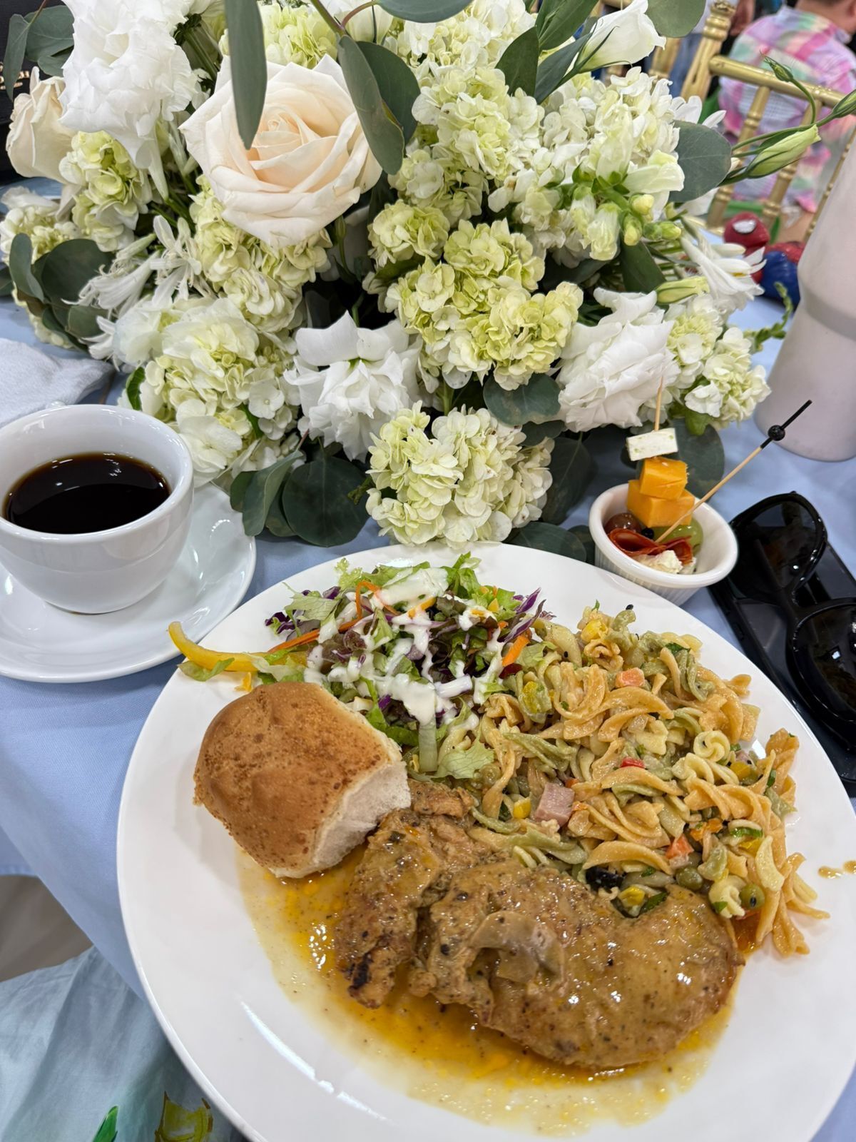 A plate with a meat dish, a salad, and a bread roll, accompanied by a cup of coffee and a bouquet of flowers.