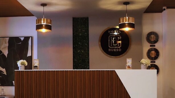 Een hotel receptiebalie met een modern ontwerp, met een houten paneel en een wit aanrecht. Daarboven hangen drie hanglampen en rechts op de muur is een klok te zien.