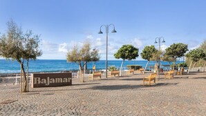 Una zona pavimentada con un letrero que dice 'Bajama', bancos, árboles y una vista despejada del mar.