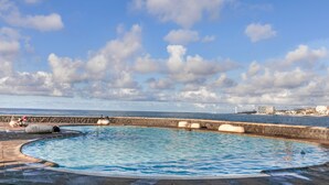 Una piscina de borde curvo, un muro de piedra y vistas al mar y edificios lejanos.