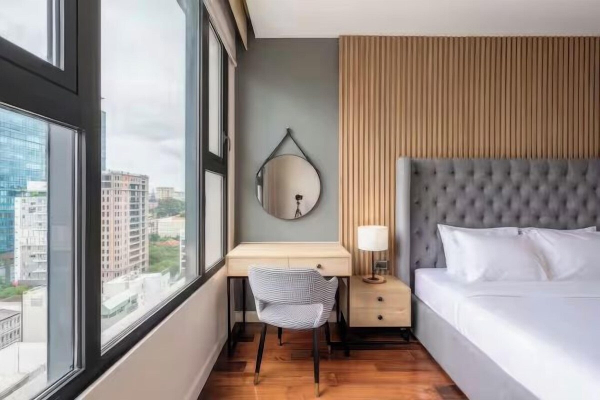 Hotellihuone, jossa on sänky, työpöytä tuolilla, pyöreä peili ja yöpöytä lampun kanssa.