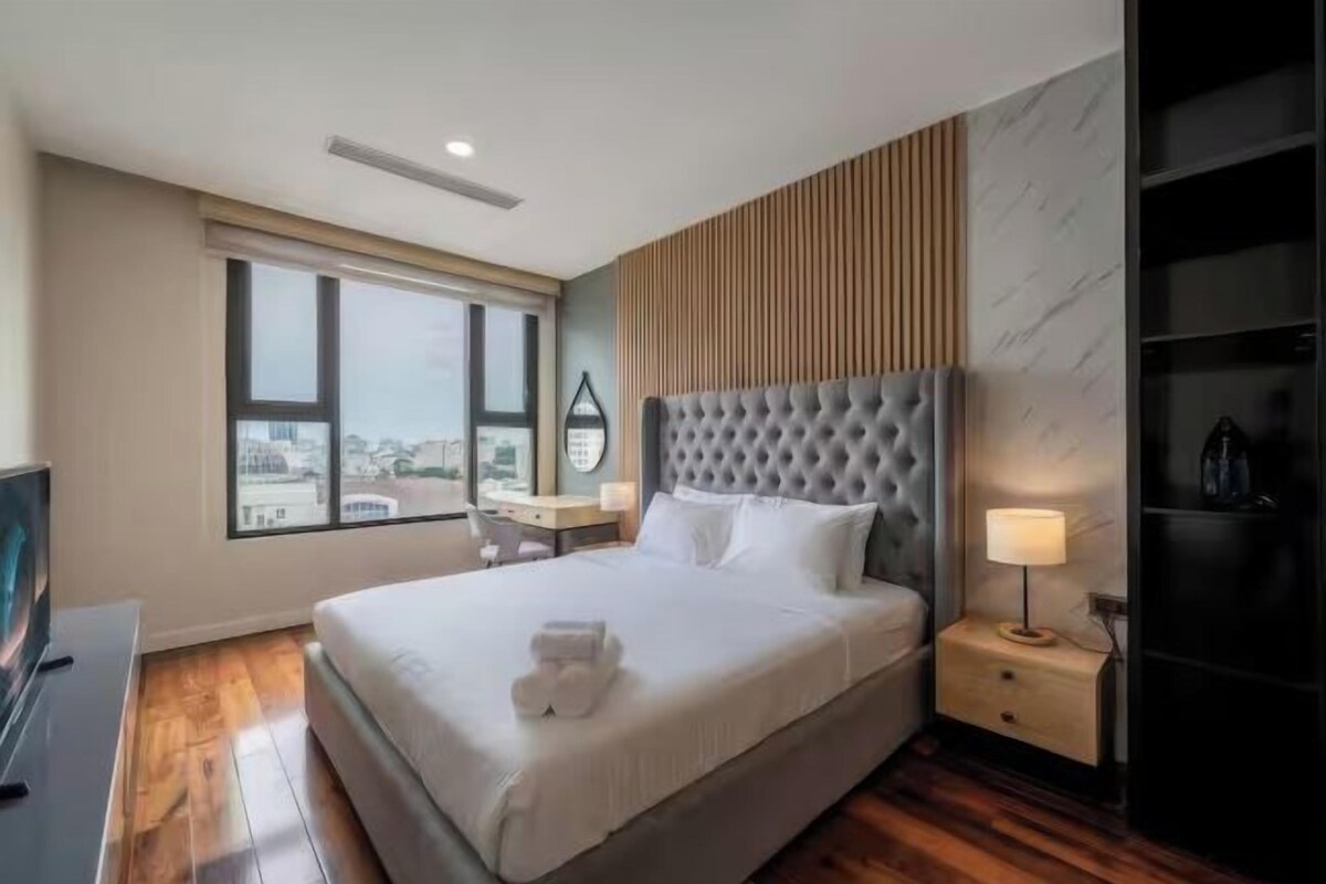 A hotel room with a large bed, a TV, a desk, and a view of the city through the window.