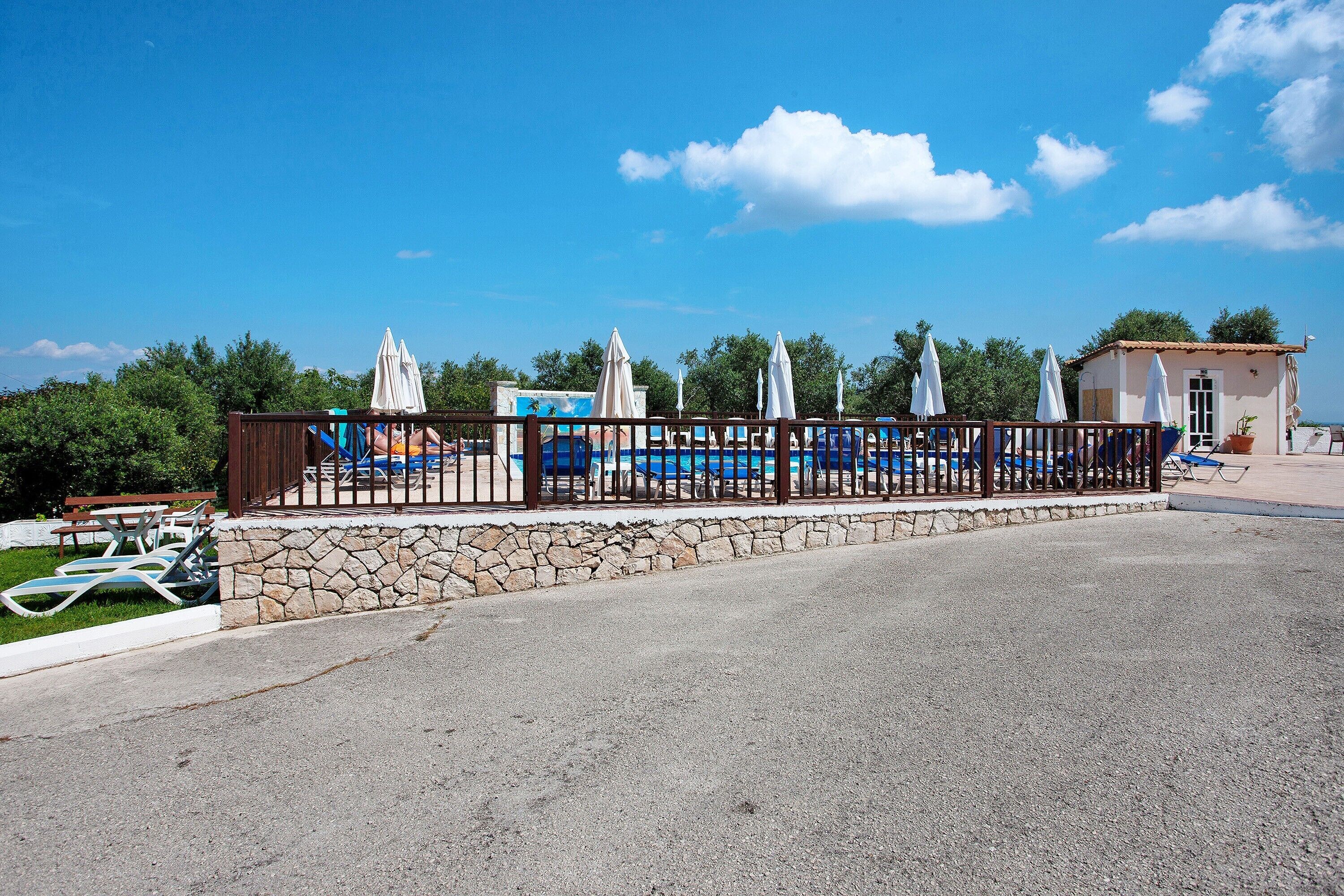 A paved area with a stone wall, a wooden fence, and a pool area with lounge chairs and umbrellas.