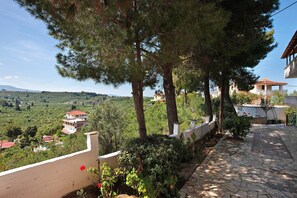 A stone pathway leading to a building with a balcony, surrounded by trees and a view of a hilly landscape.