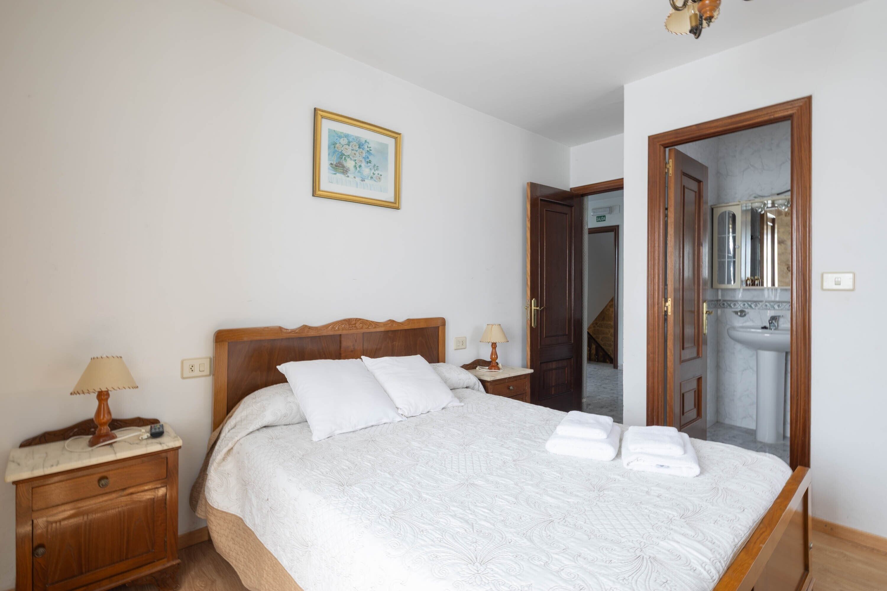 A bedroom with a bed, bedside tables, a nightstand, a framed picture, and a bathroom visible through an open door.