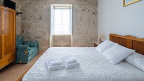 Een slaapkamer met een stenen muur, een houten garderobe, een smaragdgroene fauteuil, een bed met witte lakens en een raam met uitzicht.