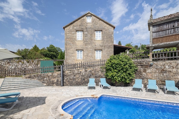 A stone building with a swimming pool, lounge chairs, and a staircase leading to a balcony.