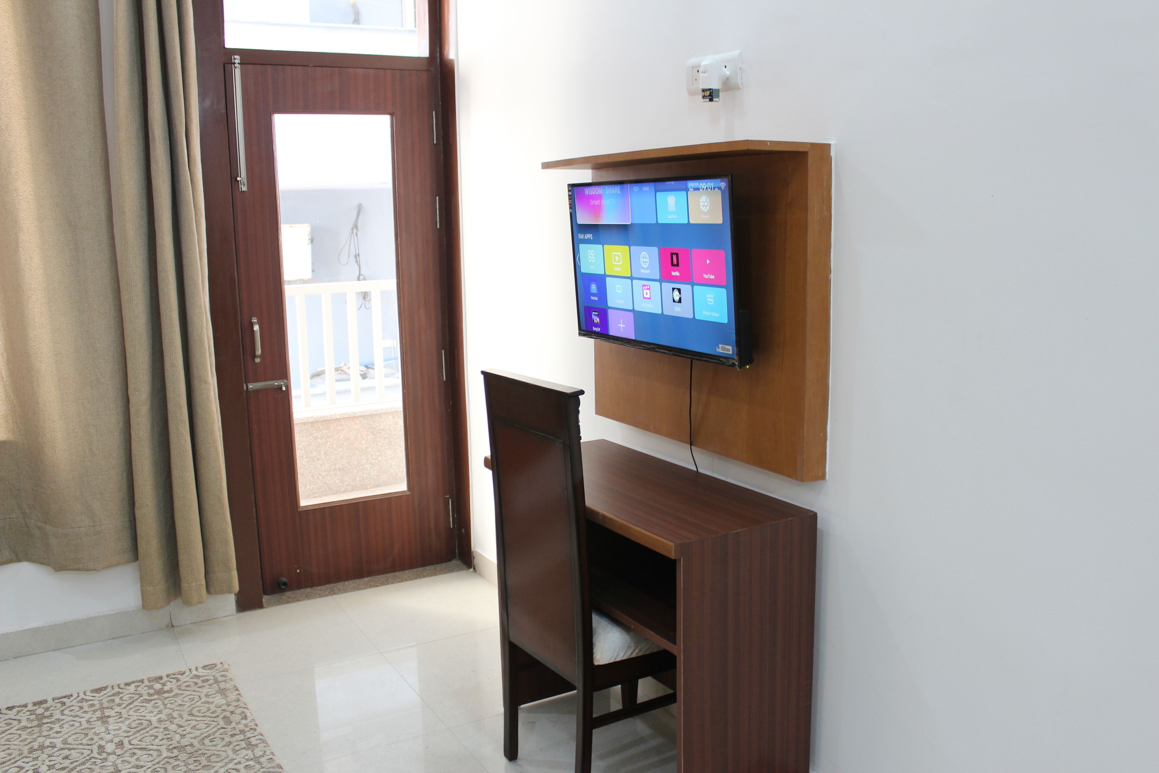 Een kamer met een houten bureau, een wandtelevisie, een deur met uitzicht en een kamerbreed tapijt met een patroon.