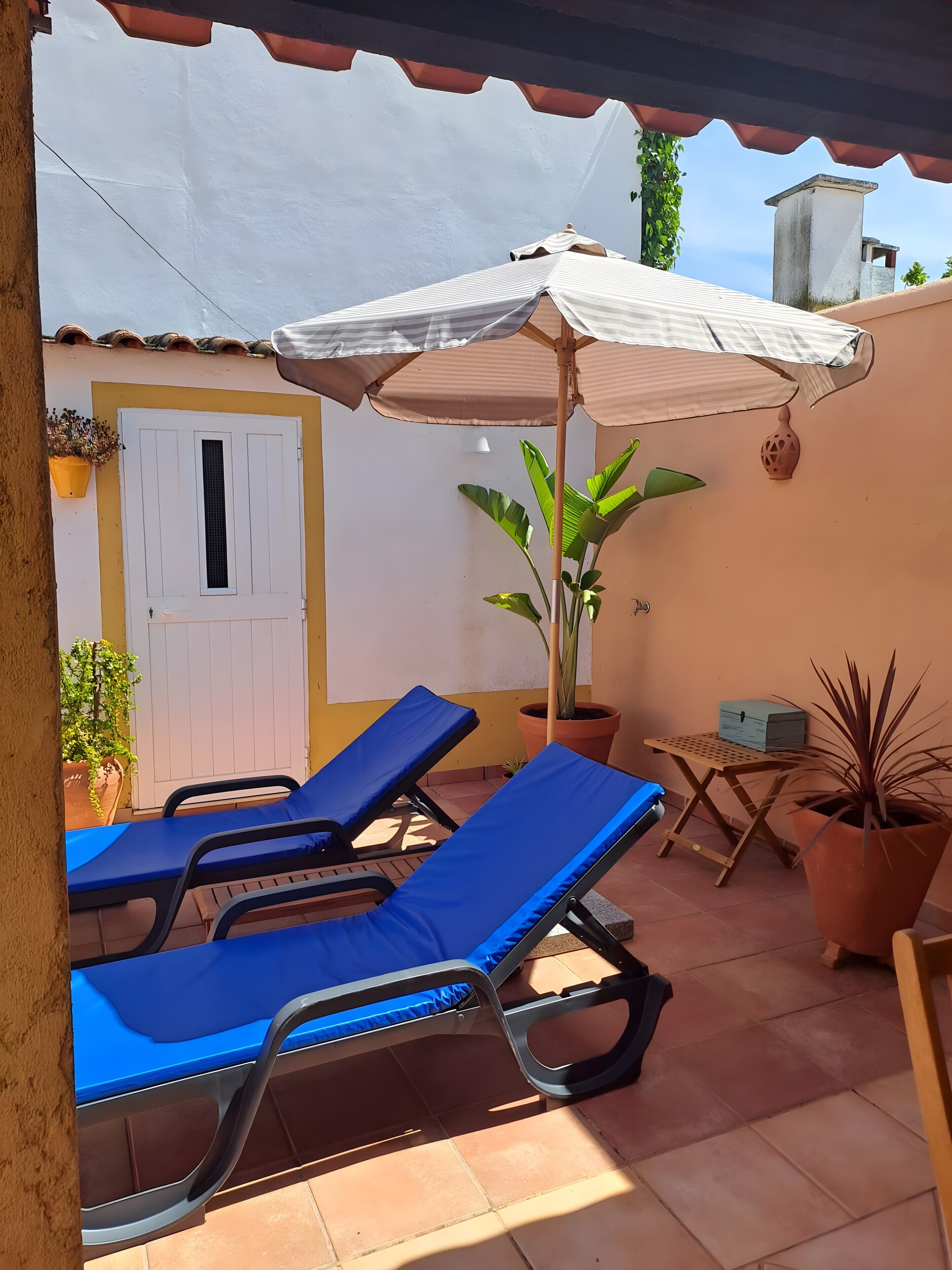 Une terrasse avec deux fauteuils bleus, une porte blanche, une plante en pot et une petite table sur laquelle se trouve une boîte.
