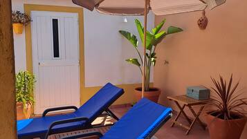 Une terrasse avec deux fauteuils bleus, une porte blanche, une plante en pot et une petite table sur laquelle se trouve une boîte.