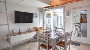 Un coin repas avec une table dressée pour quatre personnes, un téléviseur fixé au mur et une cuisine visible à travers une porte ouverte.