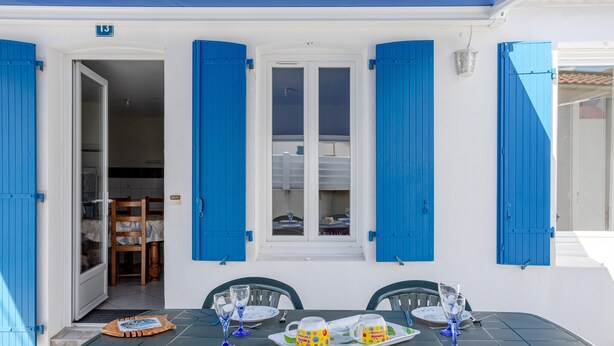 Un comedor con una mesa preparada para dos personas, una ventana con contraventanas azules y una puerta que da a un espacio interior.