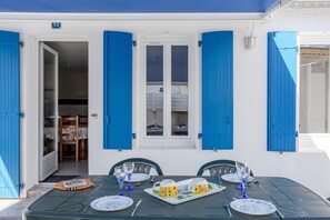 A dining area with a table set for two, a window with blue shutters, and a door leading to an interior space. - Holiday Home 'Maison Proche Océan - 6 Pers' (Bretignolles-sur-Mer)
