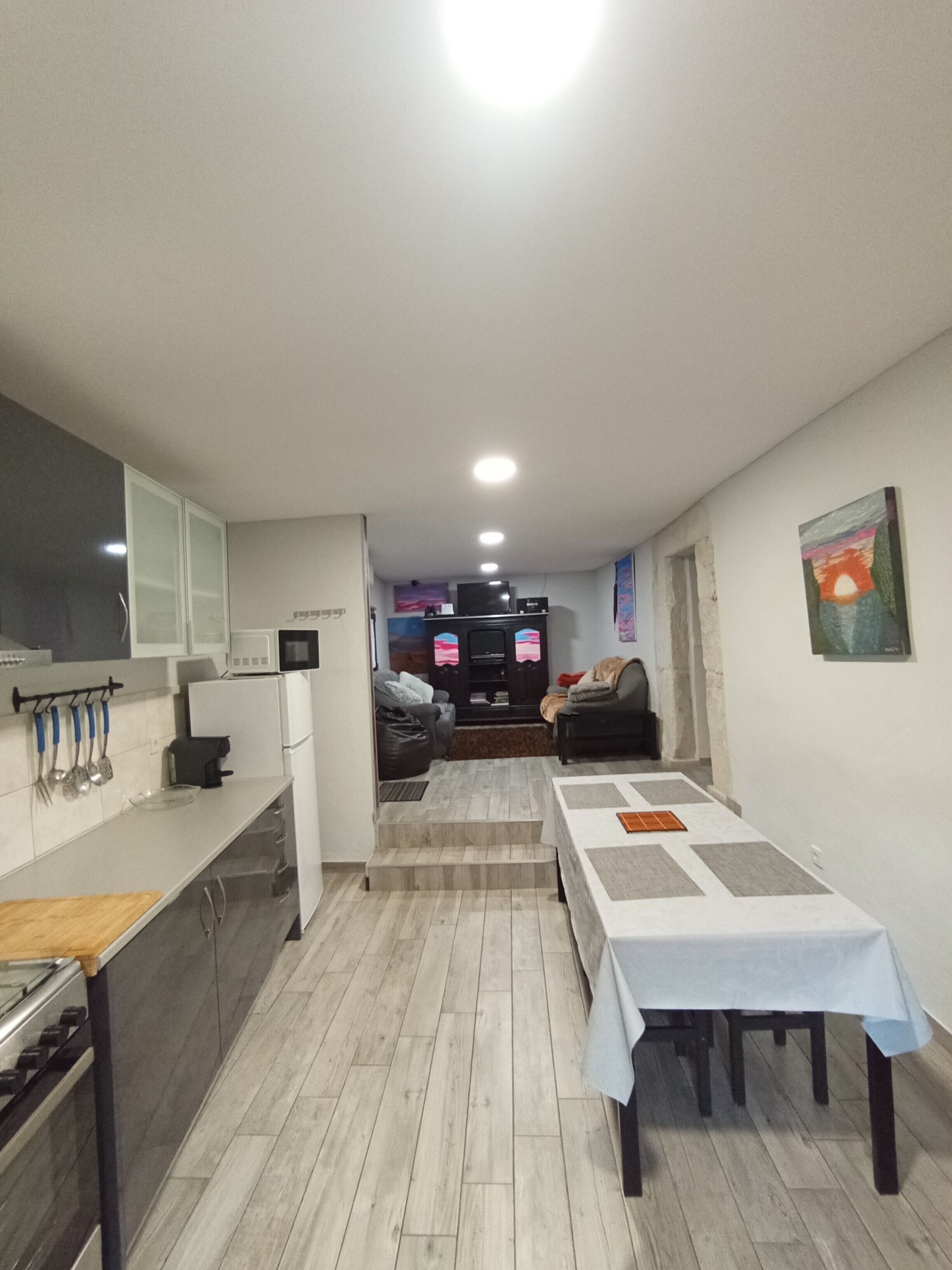 Cozinha moderna com armários brancos, área de jantar com mesa e sala de estar com sofá e televisão.