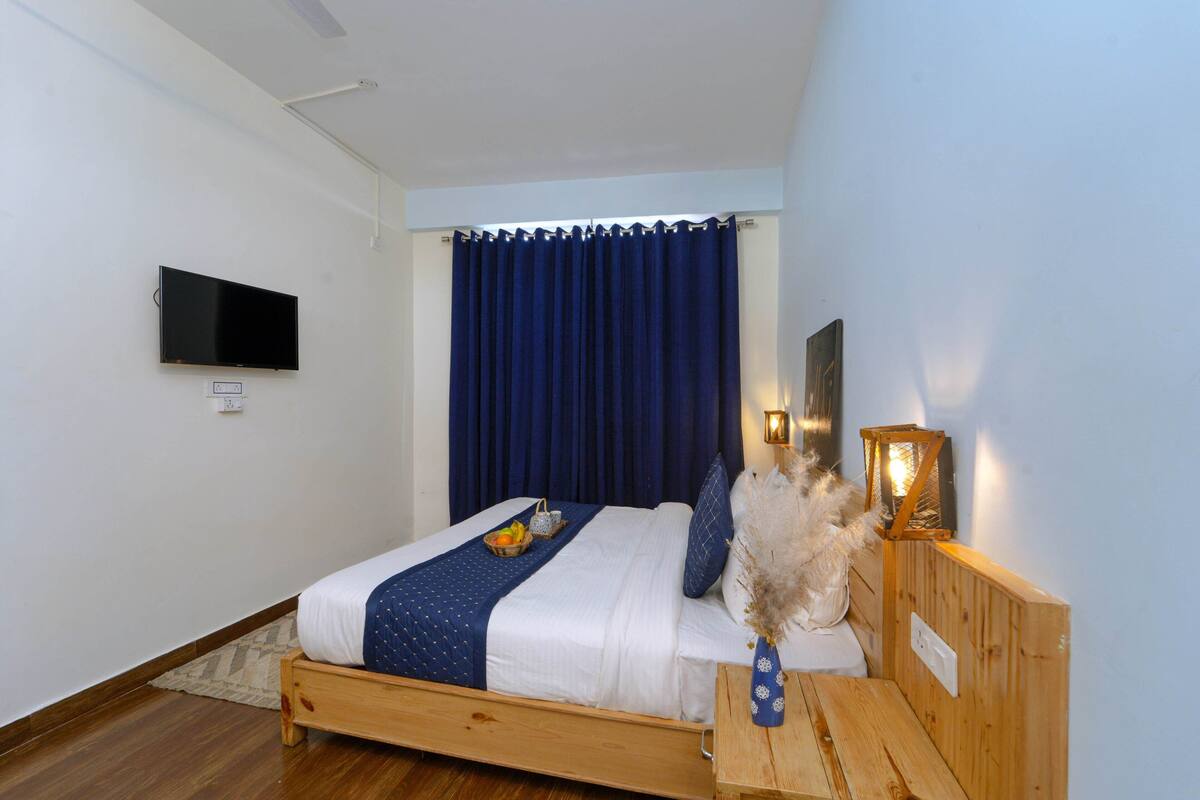 Ein Schlafzimmer mit einem hölzernen Bett, einem flachbildfernseher an der Wand, einem Nachttisch mit einer Vase und einem Fenster mit dunklen Vorhängen.