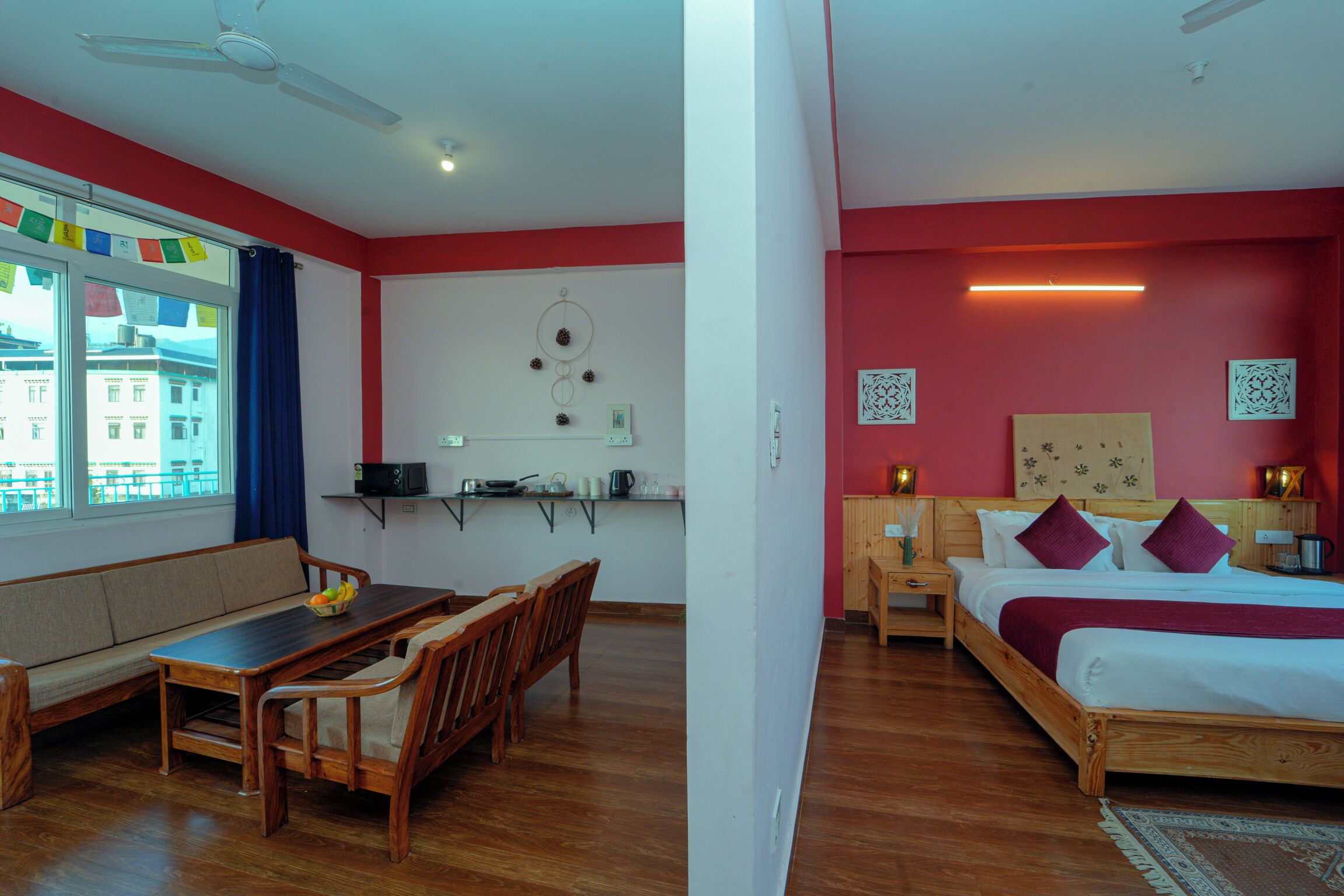 A hotel room with a wooden dining table, a sofa, a bed with a red headboard, and a view of buildings outside the window.