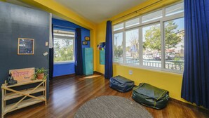 Een kamer met houten vloer, een raam met blauwe gordijnen, een bureau met een plant in pot en een mat op de vloer.