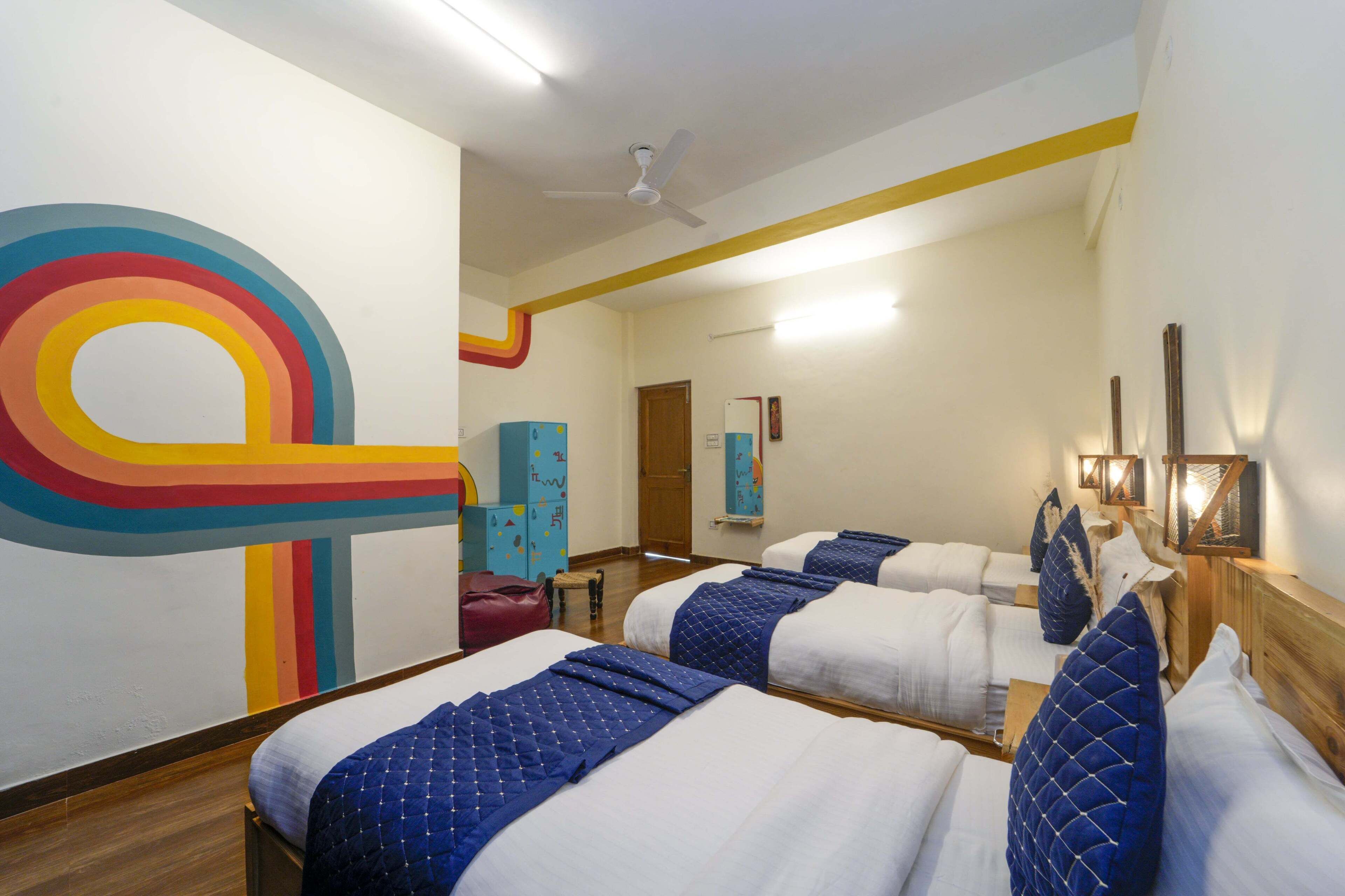 Ein Hotelzimmer mit drei Betten, einer großen abstrakten Wandkunst, einem Deckenventilator und einer Holzbank.