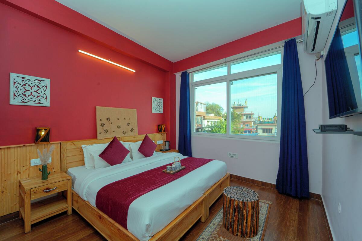 Ein Hotelzimmer mit einem großen Bett, einem Holzkopfende, roten Wänden, einem großen Fenster mit blauen Vorhängen und Blick auf Gebäude.