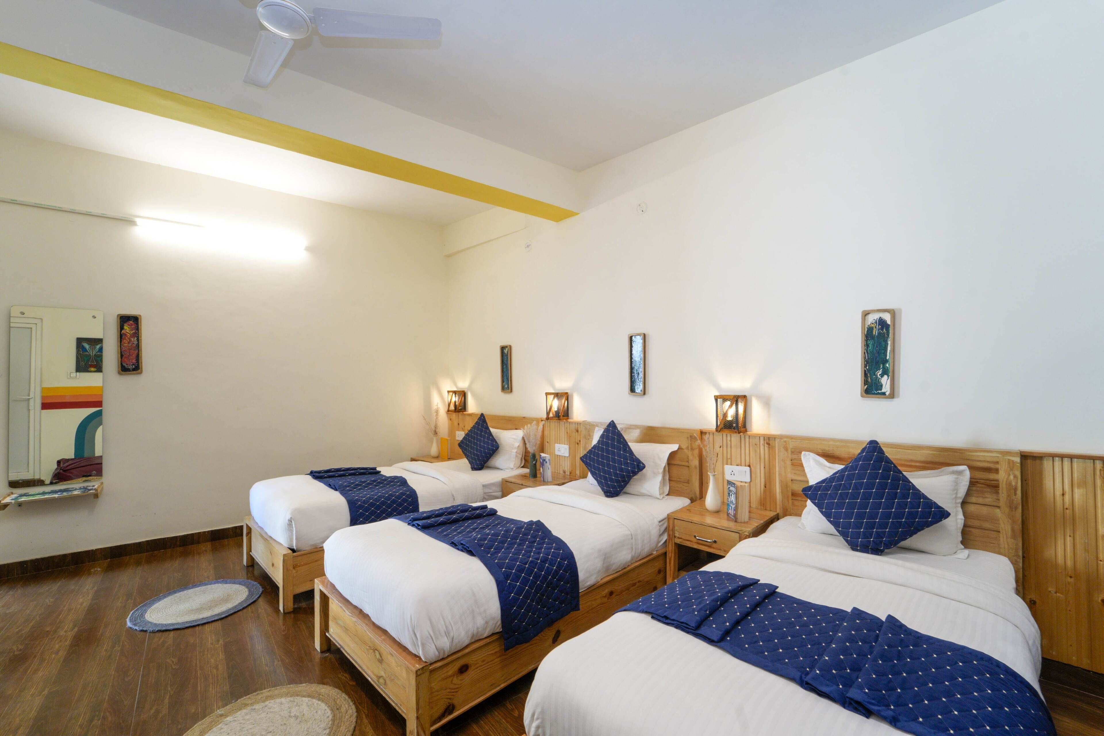 A hotel room with three wooden beds, each with white bedding and blue accents. There are bedside tables with lamps, a mirror, and a small shelf. The room has wooden flooring and a ceiling fan.