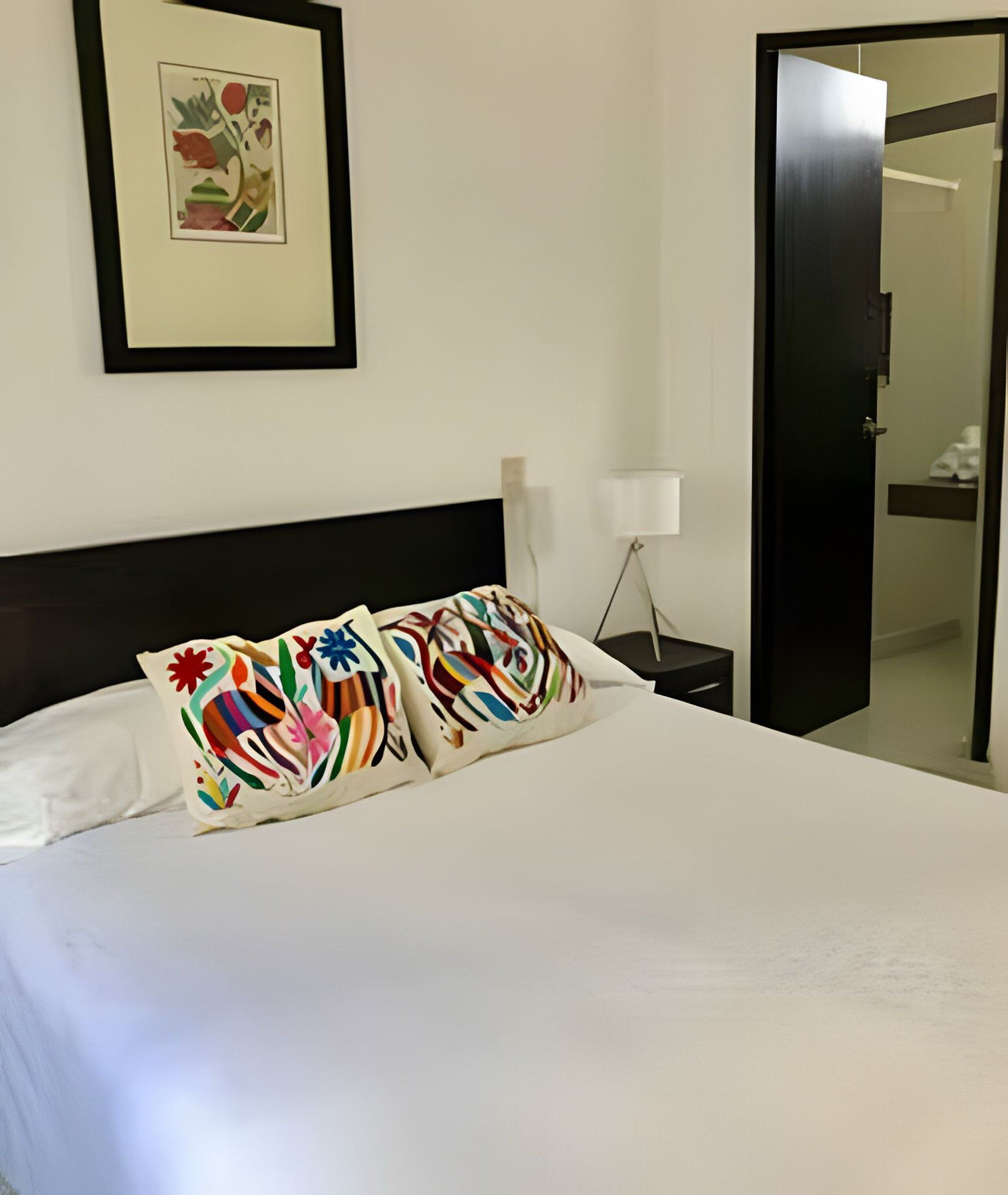 A neatly made bed with colorful pillows, a framed artwork on the wall, and a closed door to the right.
