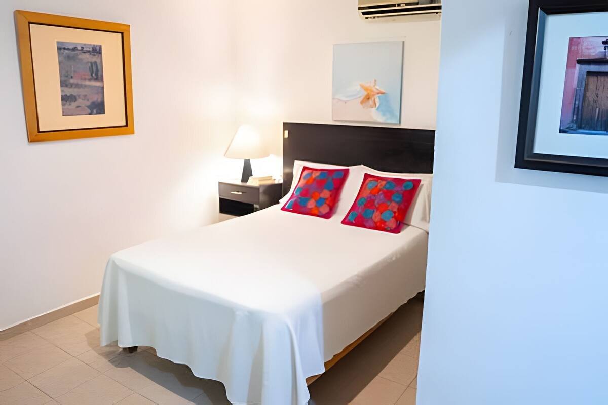 A neatly made bed with a white bedspread and colorful pillows, a bedside table with a lamp, and framed artwork on the walls.