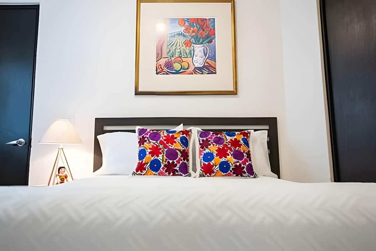A hotel room with a bed, a framed artwork above the bed, and a nightstand with a lamp.