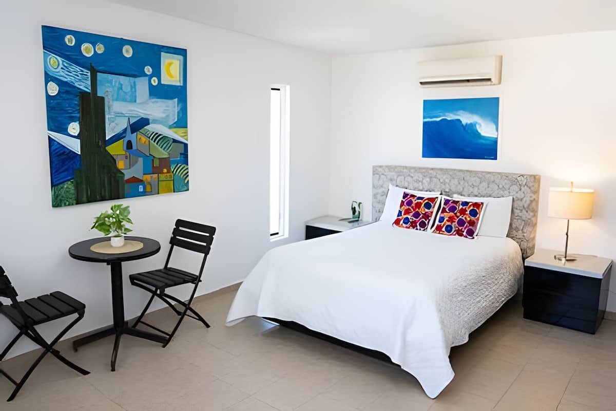 A hotel room with a bed, a chair, a small table, a plant, and artwork on the walls.