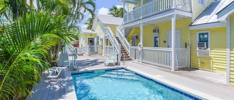 A pool area with a wooden deck, a staircase leading to a balcony, and a yellow building with white railings.