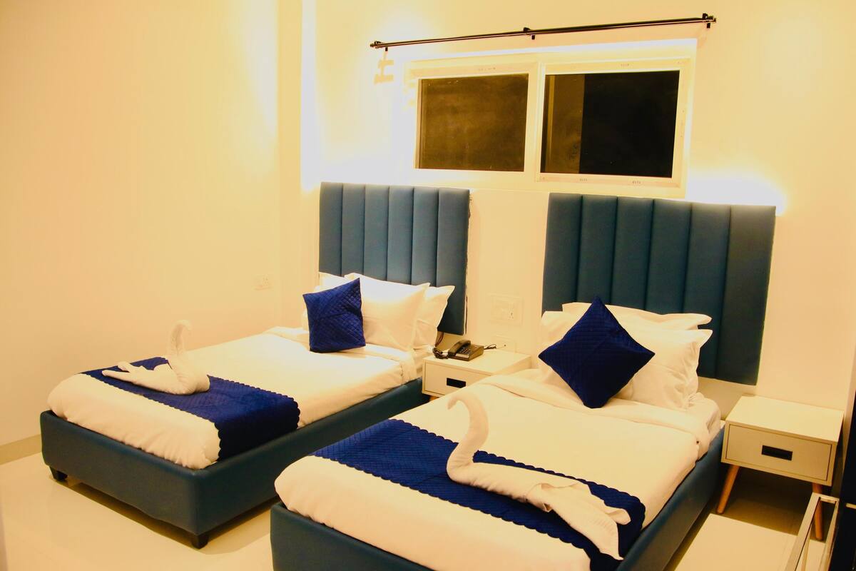 Habitación de hotel con dos camas, cada una con cabecero azul, ropa de cama blanca y manta azul. Se observa un mesita de noche con un teléfono y un cajón pequeño.