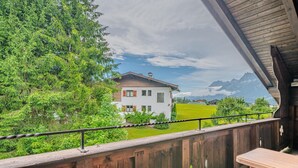Een houten balkon met uitzicht op een wit huis, een groen gazon en bergen op de achtergrond.