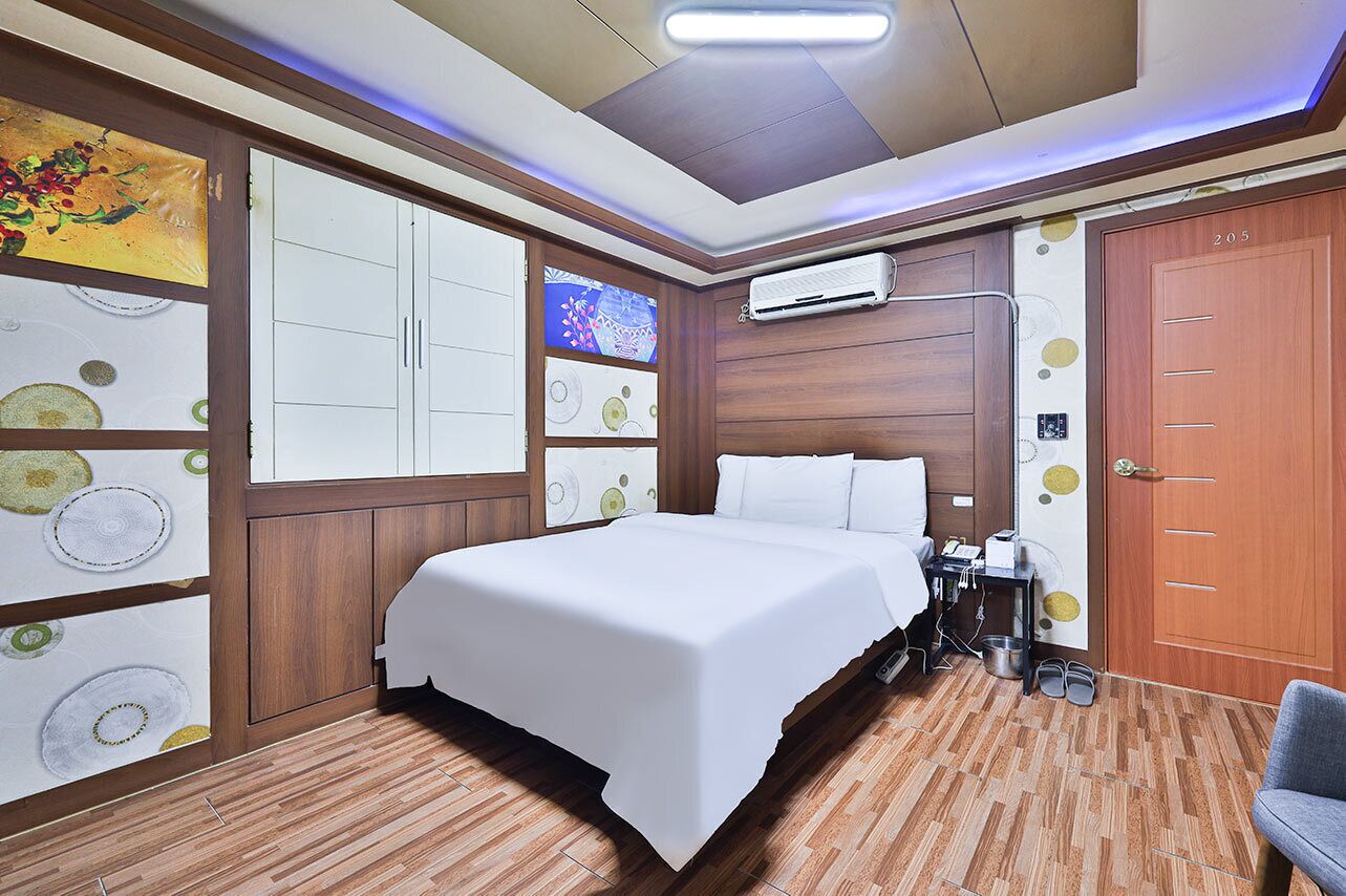 A hotel room with a bed, wooden paneling, a wardrobe, an air conditioning unit, and a door with a number.