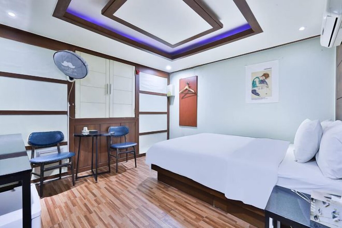 A hotel room with a bed, a desk, two chairs, a small table, a wall-mounted fan, and a picture on the wall.