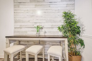 Un coin repas avec une table blanche et trois tabourets rembourrés, un vase avec des fleurs blanches et une grande plante verte dans un panier.
