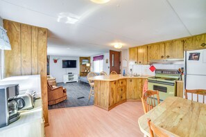 A compact kitchen with wooden cabinets, a dining area with a wooden table and chairs, and a small living space with a sofa and a television.