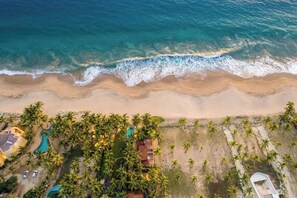 An aerial view of a beach resort with a swimming pool, palm trees, and a beach.