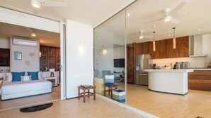 A modern hotel room with a large bed, a sitting area, and a kitchen area visible through a glass partition.