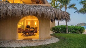 A thatched-roof structure with an interior dining area, palm trees, and a view of the sea.